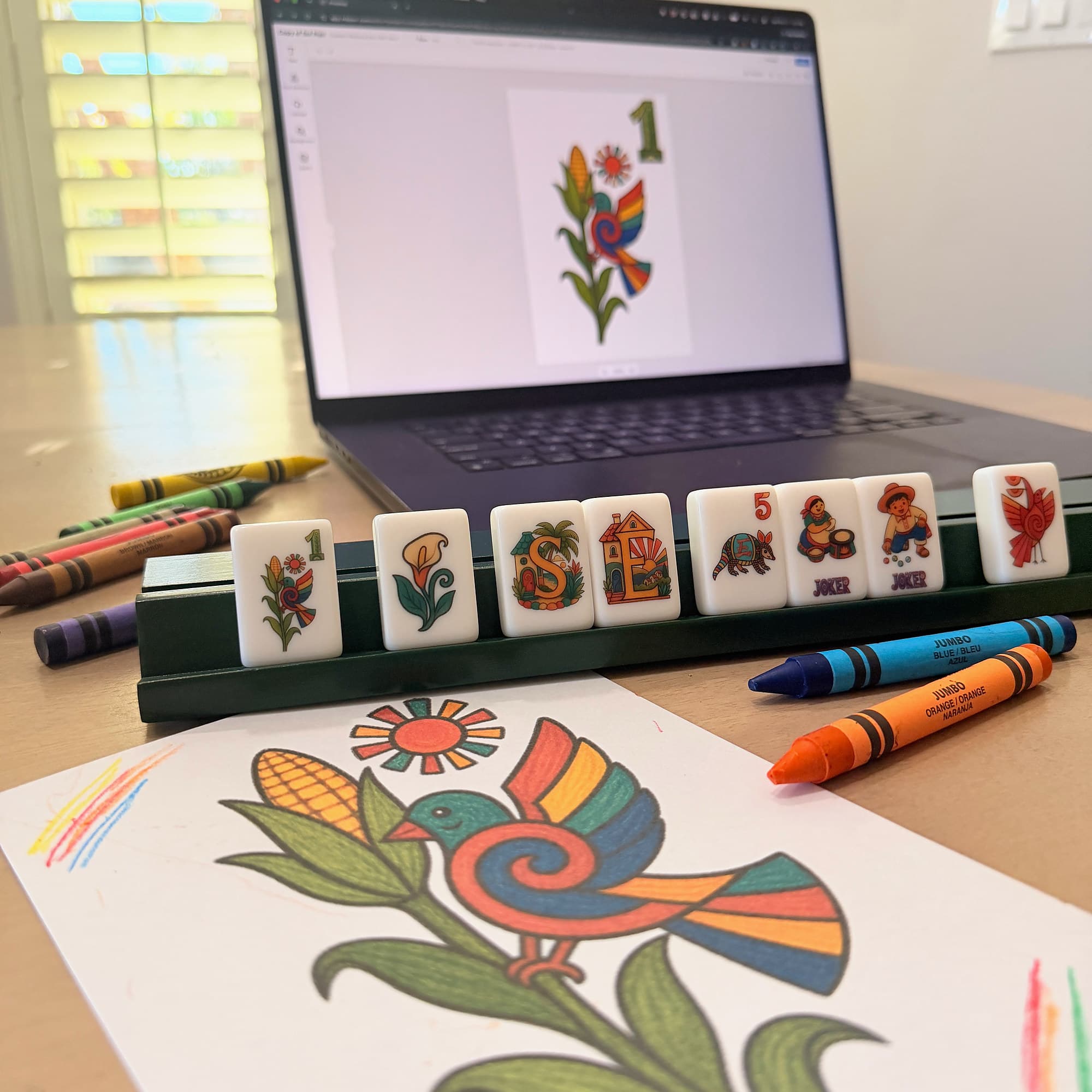 A crayon drawing of a bird on a corn stalk, a rack of printed tiles with the same bird on the 1 bam, and a laptop behind them showing that tile in the editor.